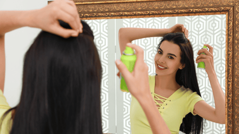 En leende kvinna med långt mörkt hår använder torrschampo framför en spegel. Hon håller en grön flaska torrschampo med sin högra hand och applicerar produkten på hårbotten medan hon lyfter sitt hår med sin vänstra hand.