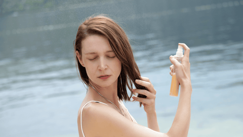 Kvinna vid vattnet sprayar värmeskydd i sitt hår på en solig dag.