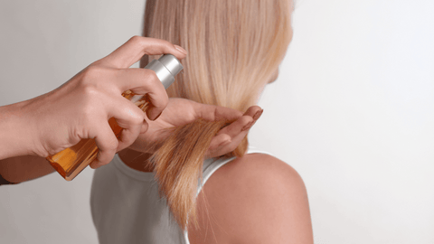 Händer som applicerar hårprodukt på blont, slätt hår mot en neutral bakgrund.