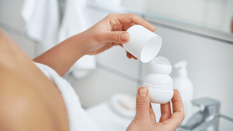 Händer som håller en öppen roll-on deodorant framför en badrumsspegel. Personen har på sig en vit frottérock och bakgrunden antyder ett ljust och rent badrum.