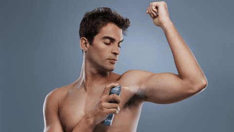En man utan tröja applicerar deodorant under armen. Han har en avslappnad hållning och ser ner på deodorantflaskan medan han håller upp sin arm för att applicera produkten. Bakgrunden är en enfärgad, neutral ton.