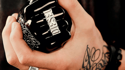 En tatuerad hand håller en svart DIESEL parfymflaska med silverbokstäver, symboliserande en modern och ungdomlig stil.