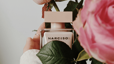 En person håller en beige parfymflaska märkt med "NARCISO", omgiven av gröna blad och en suddig rosa blomma i förgrunden, som ger en känsla av fräschhet och blommighet.
