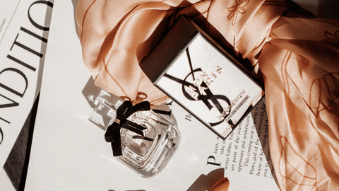 En klar parfymflaska med en svart rosett och YSL-logotyp, liggande på en öppen tidskrift och ett veckat aprikosfärgat tyg som tillför en känsla av elegans och sofistikering.