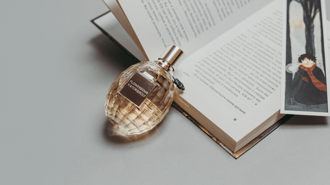 En facetterad glasflaska med guldfärgad parfym märkt 'FLOWERBOMB VIKTOR&ROLF', lutad mot en öppen bok med text och en karaktärsillustration, placerad på en neutral grå yta.