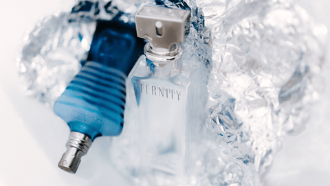 En genomskinlig parfymflaska med texten 'ETERNITY' framför en blå parfymflaska, båda på en reflekterande yta av aluminiumfolie, vilket skapar en kylig och metallisk känsla.