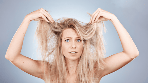 En förvånad kvinna håller upp sitt rufsiga blonda hår med båda händerna mot en lugn blå bakgrund.