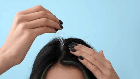 Bilden visar en person som applicerar en hårbottenbehandling med en pipett. Bakgrunden är en lugnande blå färg, vilket ger en känsla av avkoppling och vård. Personens naglar är målade i mörk färg vilket kontrasterar mot den ljusa bakgrunden.