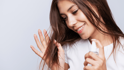En leende kvinna applicerar en sprayprodukt på sitt glänsande hår medan hon håller en flaska i den andra handen. Hon ser glad och nöjd ut medan hon tar hand om sitt hår.