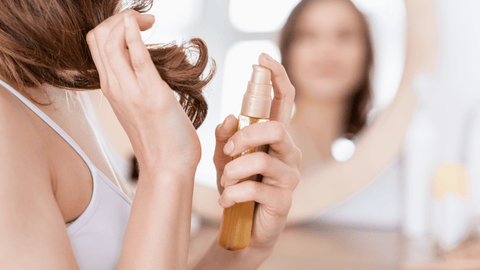 En person förbereder att applicera en produkt i håret, möjligen en hårolja eller serum. I bakgrunden återspeglas personens ansikte suddigt i en spegel. Fokus ligger på personens händer som håller en flaska med den gyllene produkten.