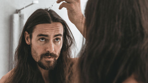 En man med långt hår applicerar en produkt, eventuellt ett hårserum eller olja, i sitt hår med hjälp av en pipett. Han ser in i spegeln med ett allvarligt uttryck medan han utför sin hårbehandlingsrutin.