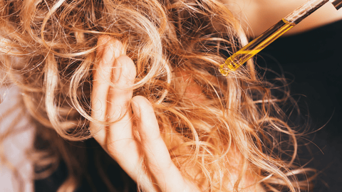 En person applicerar olja på sitt lockiga, blond hår med en pipett. Oljan syns droppa ner i håret och handen håller om en lock för att underlätta appliceringen. Detaljerna och texturen i det lockiga håret är framträdande i bilden.
