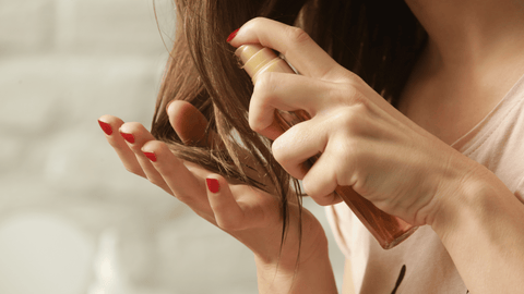 En kvinna med rödlackade naglar applicerar serum på sitt långa, bruna hår från en liten, genomskinlig flaska med brun vätska. Hon bär en ljusrosa tröja och håret ser glansigt ut. Bakgrunden är oskarp, vilket framhäver handlingen i förgrunden.