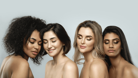 Fyra kvinnor med olika hudtoner och hårtyper poserar nära varandra. De är baraxlade och ser ut att vara i en avslappnad och förtrolig stämning mot en ljus bakgrund.