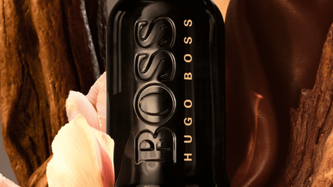 En svart parfymflaska från BOSS HUGO BOSS med en relief av logotypen, placerad mot en abstrakt bakgrund i varma bruna toner med en del av en blekrosa blomma till vänster.
