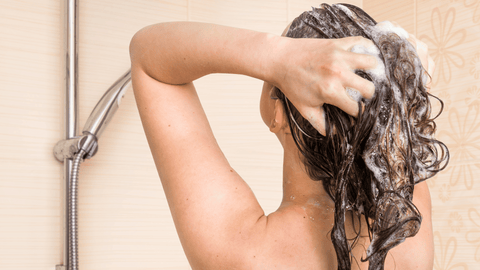 Person tvättar sitt långa, mörka hår i duschen, skummande schampo, ryggvy.