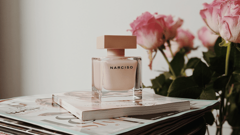 En flaska Narciso parfym i mjuk rosa ton med krämfärgad kork, placerad på en hög av magasin, med suddiga rosa rosor i bakgrunden som skapar en romantisk och sofistikerad stämning.