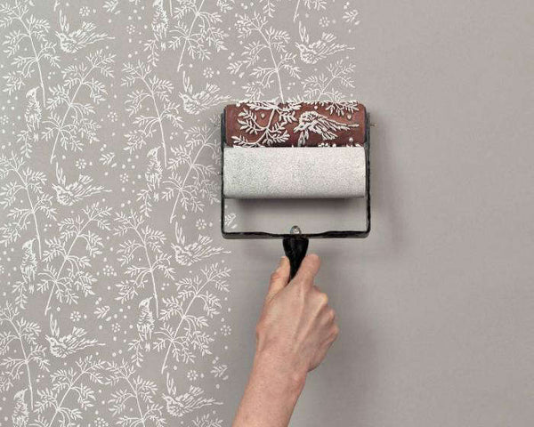 A hand applying wallpaper with a white pattern of plants and birds to a wall using a patterned paint roller