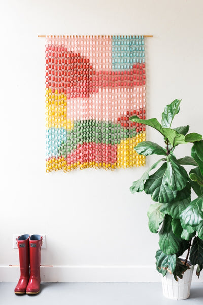 A color-coded paper tapestry on a wall. A potted plant sits on the floor as well as a pair of red rain boots