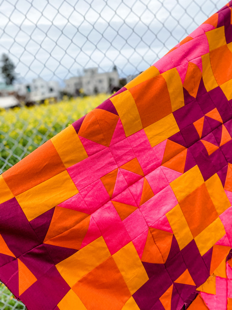 Pink and orange quilt top against a chain link fence, surrounded by a field of yellow flowers. 