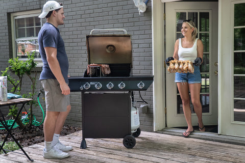 Image shows a man positioned left wearing tan shorts, white sneakers with crew socks, a blue t shirt with Blonde hair and a tan appearance smiling at a woman far right exiting a doorway holding a metal tray displaying chicken legs and other grill food items approaching a grey gas grill with the lid raised is positioned between them on a sunny day.