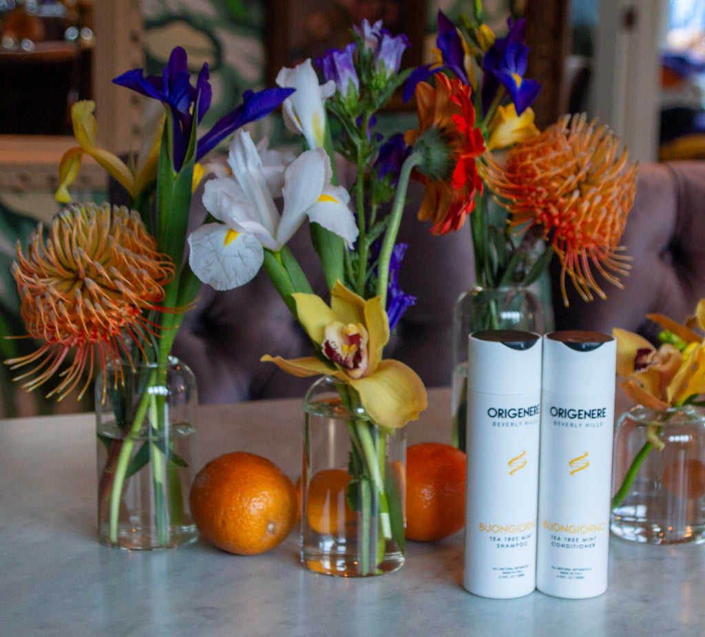 Bottles of Buongiorno Shampoo and Conditioner on a marble table surrounded by beakers of flowers and citrus fruits, with oranges next to them