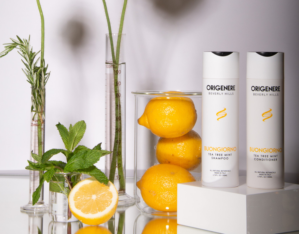 Origenere's Buongiorno Shampoo and Conditioner on a table next to a beaker of lemons, beaker of mint, a test tube of rosemary and a test tube with flowersa