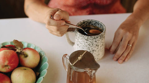 Hand spooning honey into tea with a bowl of apples nearby