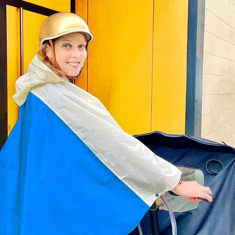 Blauer Regenponcho für Damen auf dem Fahrrad mit Helm und Kapuze. Die Frau steht vor einem gelben Hintergrund und schaut lächelnd den Betrachter an. Lastenrad Regencape