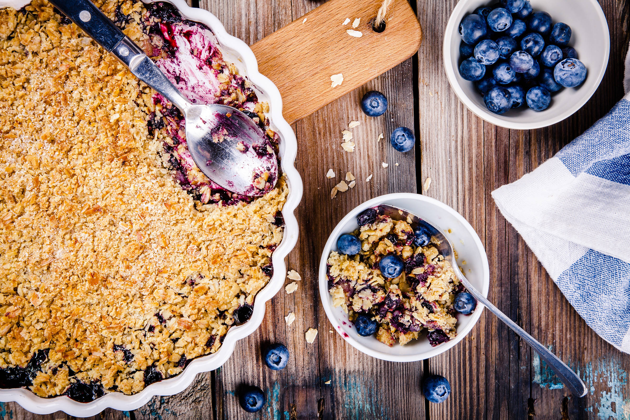 Blueberry crumble in a pie dish on wooden board with a serving in a dessert bowl served with fresh blueberries.