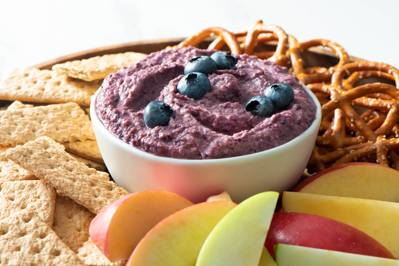 Blueberry hummus in a bowl topped with fresh blueberries, alongside crackers, fresh fruit, and pretzels.