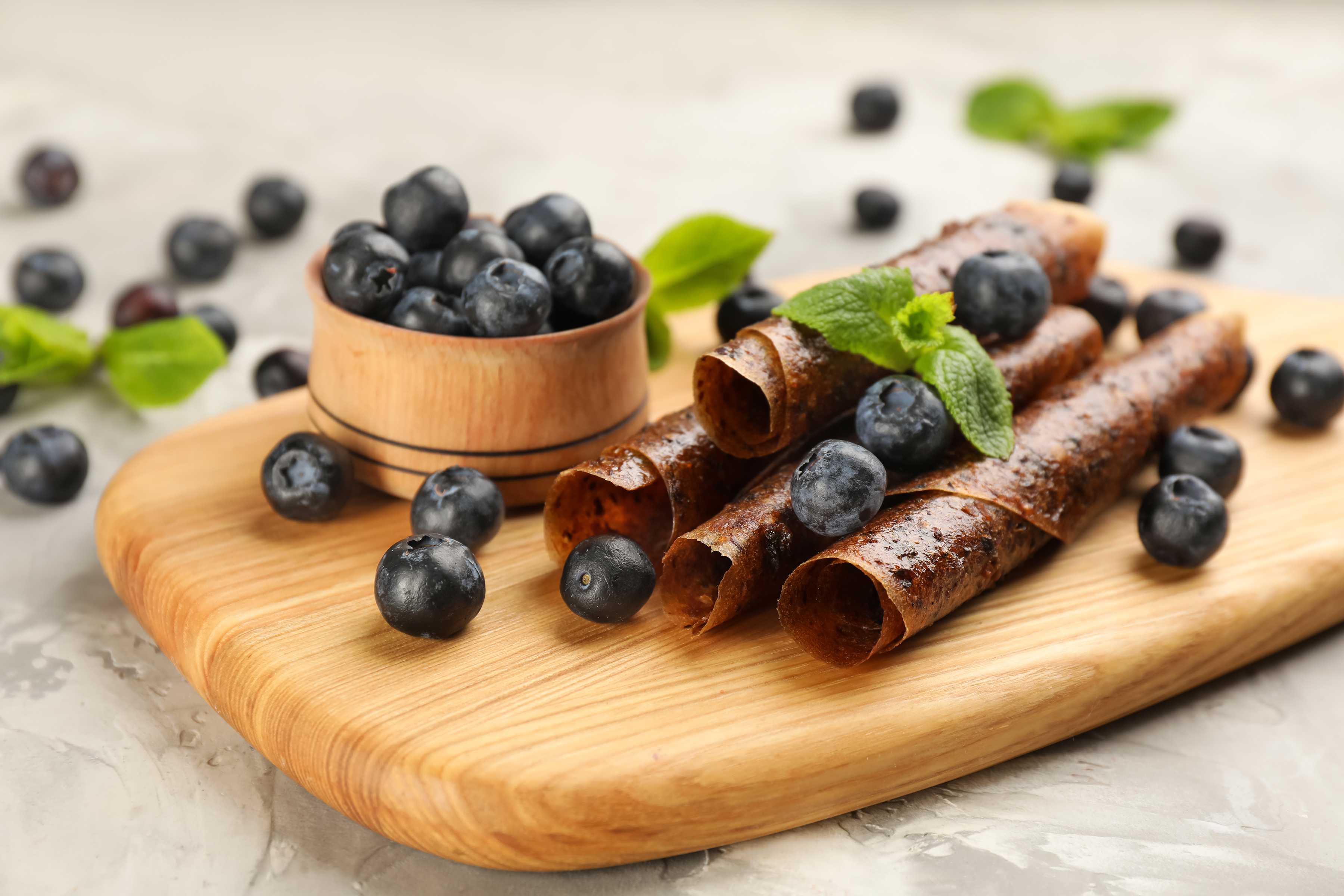 Rolls of blueberry fruit leather presented on a wooden serving board with fresh blueberries.