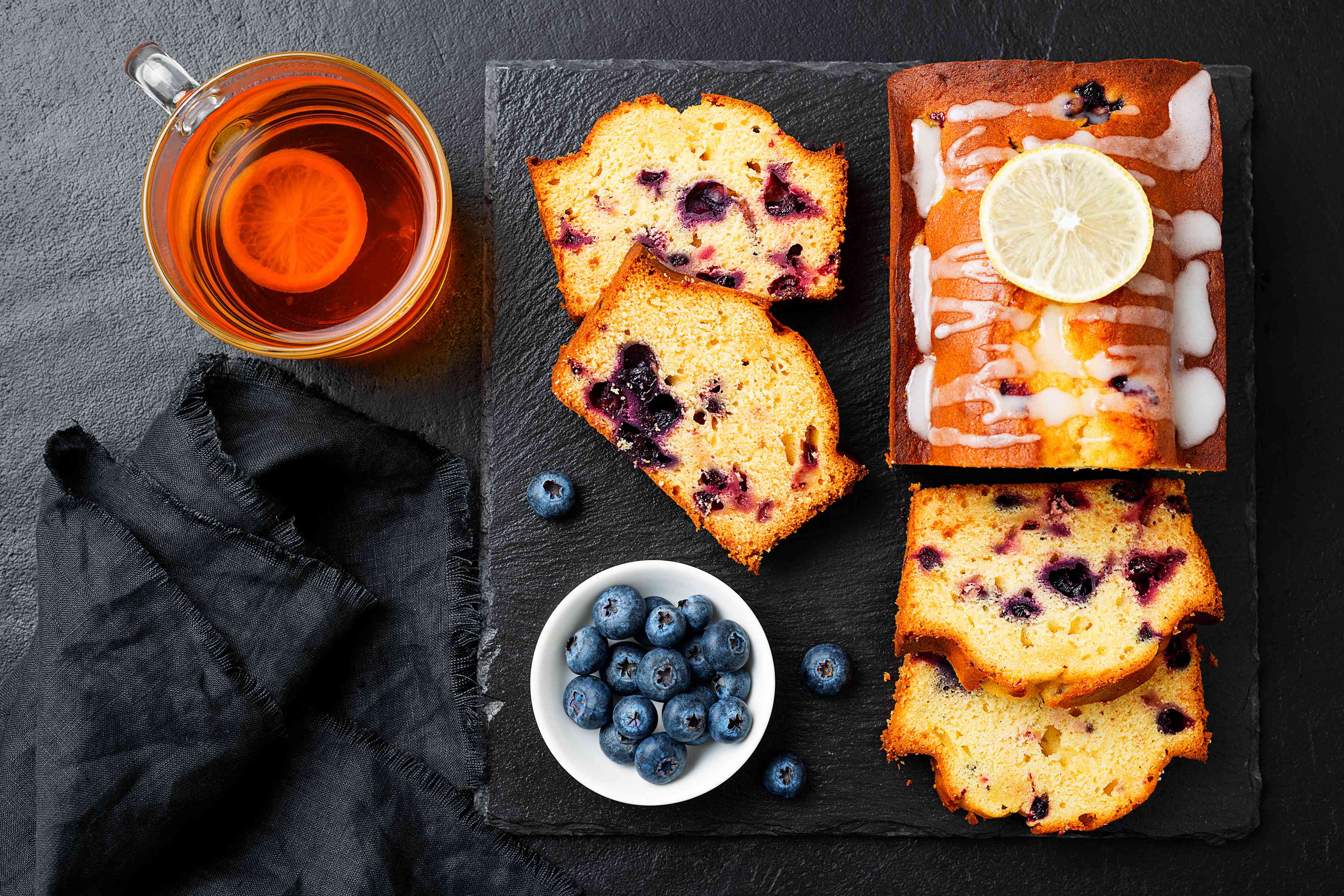 Vegan blueberry lemon cake presented on a black serving board with a bowl of blueberries, alongside a cup of herbal tea.