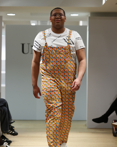 Wesley, a black man with autism and vitiligo, is walking the runway at London Fashion Week. He is wearing Lucy and Yak / Unhidden collaboration dungerees and a "Disabled is not a bad word" white tshirt.