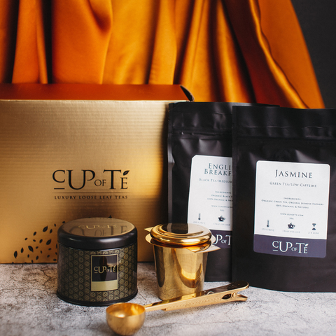 A product image of two packages of tea, a box with "Cup of Te" written on it, a gold spoon, tea infuser and gold tin. The products are placed on a marble counter. The background has orange drapes.