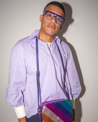 Lee Chambers is a male presenting person. He is wearing a white and purple checkered shirt., a rainbow cross body bag and eye glasses with a blue rim. He is standing in front of a cream with his hands in his pockets.