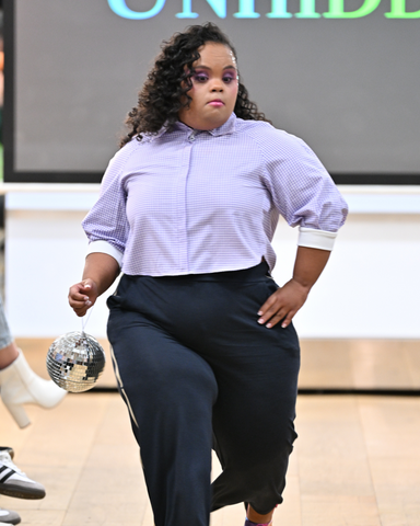 Elena, a mixed heritage woman with Downs syndrome, is walking down the runway. She is wearing a purple gingham shirt with navy trousers. She has her hand on her hip and is carrying a sparkly disco ball in the other hand.