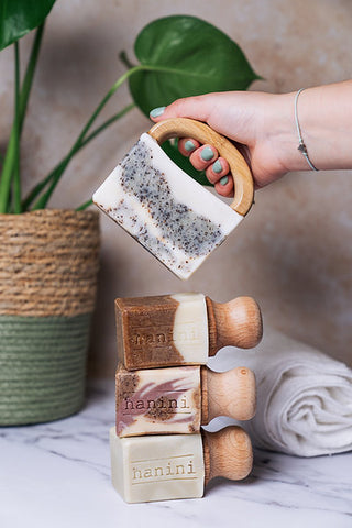 In this image, three bars of soaps with wooden top handles are placed on top of each other, embellished with the name of the brand "hanini". The soaps are in different shades of brown and are placed on a marble counter. A hand holding a bar of soap by a wooden hand attached to it seen right above the stack of soap. The hand has nails painted in bright light blue nail varnish. In the background of the counter is a potted plant on the left hand side and on the right hand side is a rolled up towel.