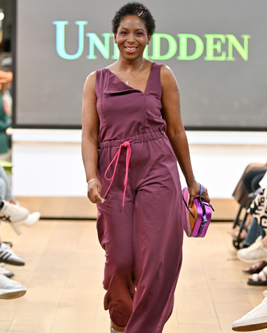 Jacqueline is a black woman with short hair. She is wearing a purple jumpsuit and is walking the runway with a bright smile while holding a purple purse.