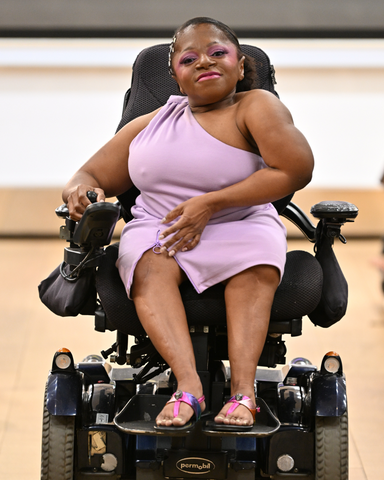 Monique is a short statured, black woman with slicked back black hair. She is wearing a lavender colored one shoulder dress, rainbow sandals and is on a power chair. She is on the Unhidden runway.