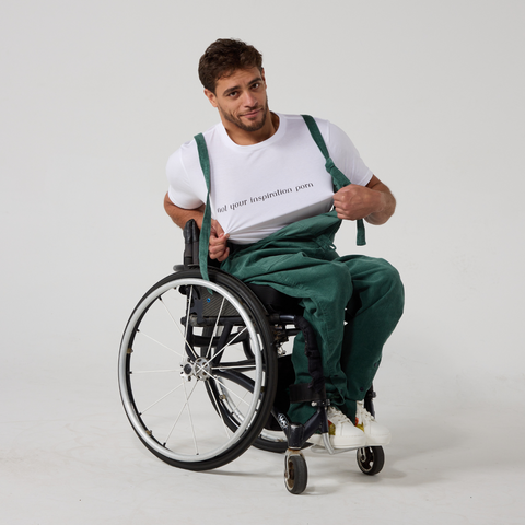 Moatez, a Tunisian man, is wearing a green corduroy dungaree and a white t-shirt with text across the chest reading "not your inspiration porn". He is stretching the shirt towards the camera. He is sitting on a manual wheelchair. The background is cream in color.