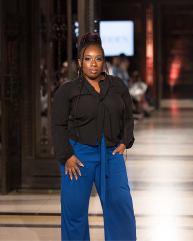 Glynis, black woman with an arm limb difference. She is wearing a black blouse and blue trousers. She has diamantes on her hair parting. She is on the runway and one of her hands are in her pockets while the other is resting on her thigh.