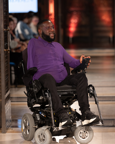 Adrian, a male presenting man in a power chair. He is dressed in purple shirt and black trousers. He is on the runway and is posing sideways.