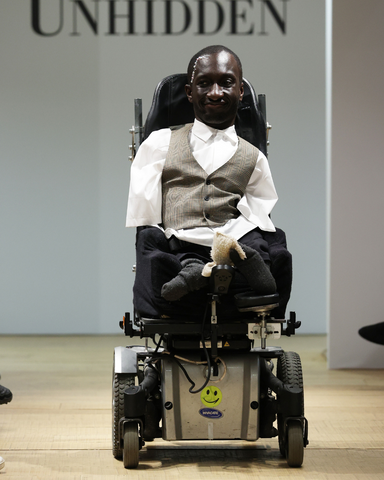 Isaac, a black man, has white pearls decorating his face and close cut hair. He is wearing a white short sleeve shirt under a prince of wales check waistcoat with black shorts. He has grey socks on and is using a power chair. Behind them is a white back drop with Unhidden in black text visible above his head.