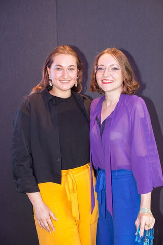 Rachael, a white woman with long brown hair wearing a black shirt and yellow jersey culottes is standing with Victoria, a white woman with mid length brown hair wearing a purple pussy bow shirt and blue jersey culottes and holding a blue mobility aide. They are both smiling at the camera.