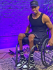 A Black man called Antwan Tolliver sits in a manual wheelchair. He is wearing a black vest and sports shorts. The wall behind him is bright purple fading to pink at the floor. He is also wearing a black cap.