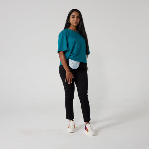 Tanya is a south asian woman who is posing with a smile, facing the camera. She is dressed in a teal top, black trousers and a silver sparkly stoma bag. The background is cream.