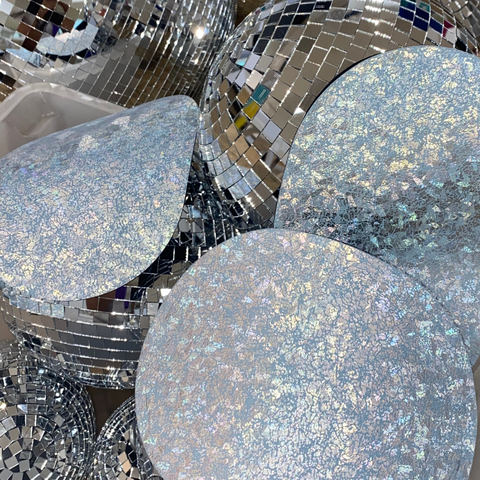 3 Stoma bag covers with silver holographic print are placed on disco balls. The stoma bag covers are from the brand Ostique.
