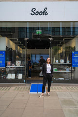 Victoria stands at the shop front of Sook Oxford Street. There is a blue wheelchair ramp leading in to the shop. Victoria is wearing black trousers and jacket and a white 'Unhidden' T-shirt.