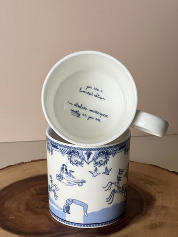 A mug is placed horizontally on an another mug in way that the bottom on the top mug is displayed to the camera. The inside of mug reads "you are a limited edition. an absolute masterpiece exactly as you are." The mug is white, the writing and patterns are blue.